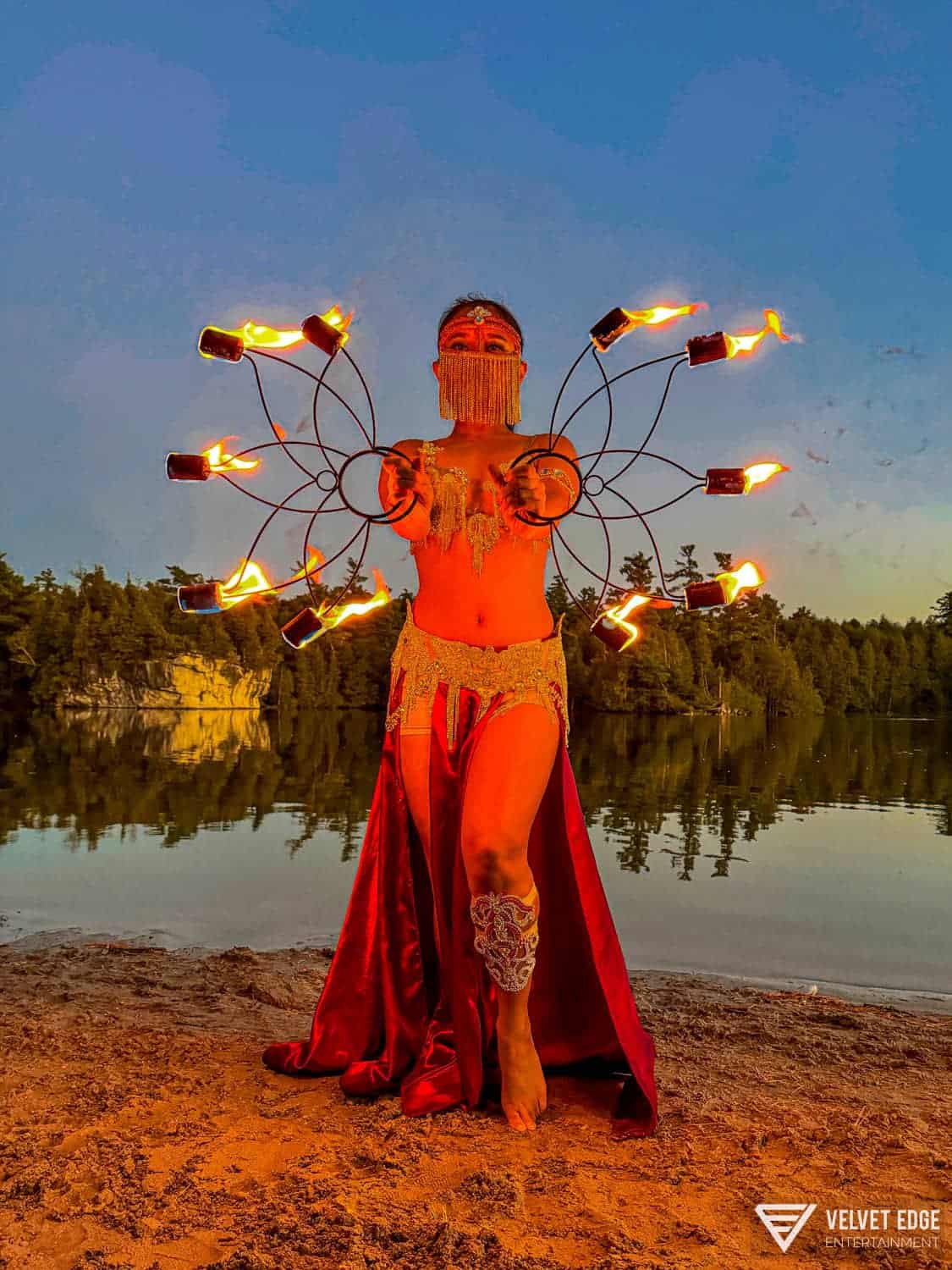 Vibrant fire dancer in elaborate costume performing at sunset on lakeside.